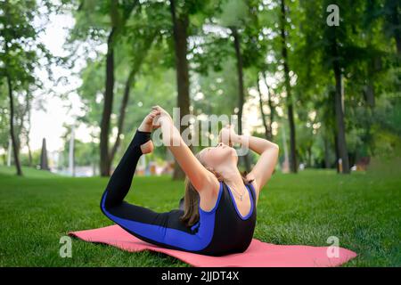 dans le parc, une petite fille se plie en arrière, embrassant ses jambes, exécute des éléments de yoga, fait des étirements Banque D'Images