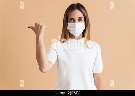 Blanc femme moyenne dans le masque de visage pointant le doigt de côté isolé sur fond beige Banque D'Images