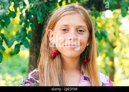 Refocalisation belle blonde jeune fille avec cerise douce sur l'oreille. Belle adolescente souriante à proximité, contre le vert du parc d'été. Boucles d'oreilles pour enfants. Somme Banque D'Images