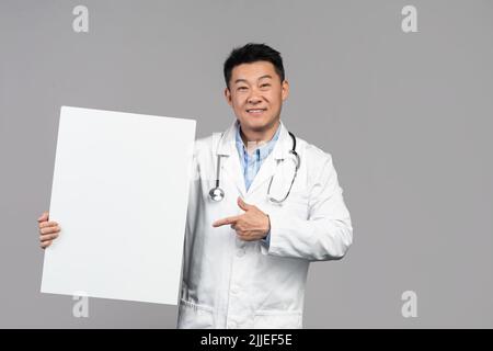 Souriant beau adulte asiatique homme médecin en manteau blanc avec stéthoscope pointe le doigt sur la bannière Banque D'Images