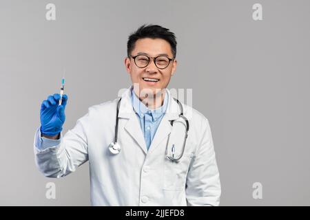 Joyeux homme japonais mature médecin en manteau blanc, verres, gants de protection tiennent la seringue Banque D'Images