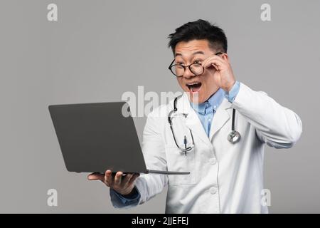 Choqué gai millénaire asiatique homme docteur en manteau blanc avec la bouche ouverte prend des lunettes, regarde l'ordinateur portable Banque D'Images