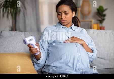 Femme noire enceinte malade ayant de la fièvre tenant le thermomètre à la maison Banque D'Images