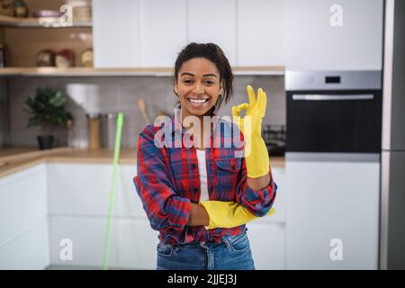 Femme au foyer noire millénaire gaie dans des gants en caoutchouc montrant le signe ok avec la main Banque D'Images