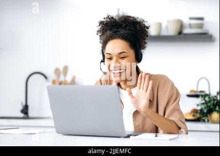 Une jeune femme afro-américaine heureuse, qui se sert d'un micro-casque et d'un ordinateur portable, parle lors d'une vidéoconférence avec un client ou un collègue assis dans la cuisine à la maison, souriant et amical Banque D'Images