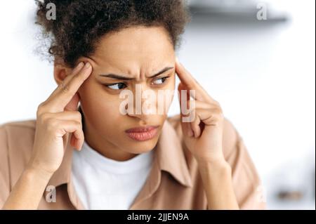 Gros plan de femme fatiguée afro-américaine stressante, fatiguée du travail, surtravaille, souffrant de maux de tête, migraine, massages de ses temples, regarde sur le côté, a besoin de repos Banque D'Images