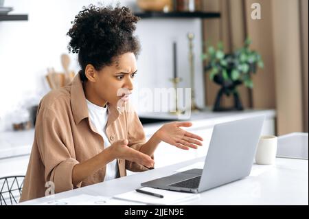 Une jeune femme brunette afro-américaine stupéfaite, pigiste, Manager, assis à la table dans la cuisine, utilise un ordinateur portable tandis que le travail à distance, choqué par les nouvelles ou le message, regardant l'écran confus Banque D'Images