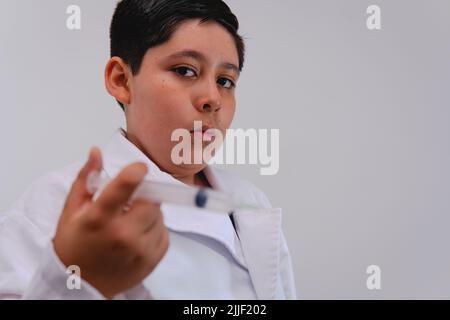 Un garçon habillé comme un médecin dans un manteau blanc et avec une seringue dans sa main. Petit garçon habillé comme un médecin posant avec une seringue comme s'il allait donner une injection. Banque D'Images