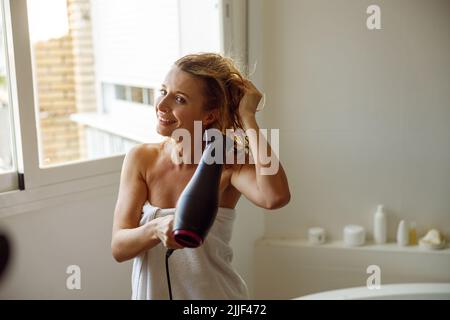 Gai jeune caucasien belle femme séchant les cheveux avec sèche-cheveux après avoir pris la douche. Banque D'Images