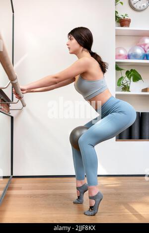 Vue latérale complète de la ballerine FIT en mode actif, squat sur barre avec ballon pendant la répétition en studio léger Banque D'Images