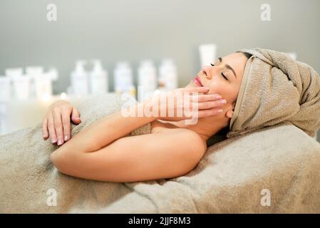 Vue latérale d'une femme détendue enveloppée d'une serviette touchant la joue en étant allongé sur le canapé pendant la procédure de spa dans le salon Banque D'Images