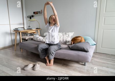 Paisible femme d'âge moyen vivant seule avec un chien assis avec les yeux fermés et les bras levés sur le lit Banque D'Images