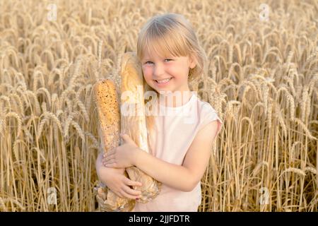 Bonne petite fille blonde tenant une baguette de pain française dans un champ de blé. Enfants avec du pain. Récolte de blé Banque D'Images