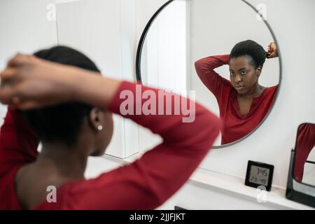 La routine de deuil de la femme africaine noire fixant ses cheveux et regardant dans le miroir de salle de bains. Coiffage et préparation Banque D'Images