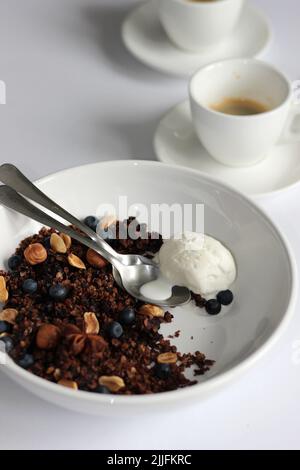 Dessert au chocolat et à la granola aux myrtilles, aux noix et à la cuillère de glace à la vanille sur fond de table blanc avec café. Banque D'Images