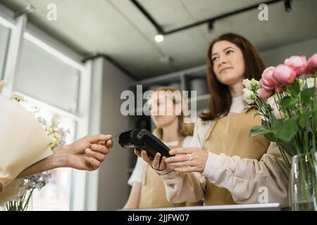 Deux femmes souriantes fleuristes tenant la machine de lecteur de carte au comptoir avec le client payant avec la carte de crédit. Jeune fleuriste assistant de boutique tenant la machine de paiement tandis que l'acheteur achète un bouquet de fleurs. Banque D'Images