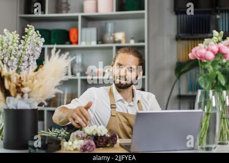 Portrait d'un fleuriste attrayant travaillant sur un ordinateur sur le lieu de travail. Homme barbu concentré assis au comptoir dans un magasin floral et dactylographiant sur un ordinateur portable. Banque D'Images