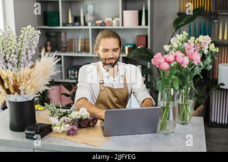 Portrait d'un fleuriste attrayant travaillant sur un ordinateur sur le lieu de travail. Homme barbu concentré assis au comptoir dans un magasin floral et dactylographiant sur un ordinateur portable. Banque D'Images