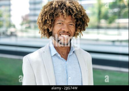 Gros plan d'un bel homme afro-américain ou brésilien positif aux cheveux bouclés, habillé en tenue habillée élégante, debout à l'extérieur, regardant l'appareil photo, souriant amical Banque D'Images