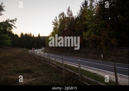 Une belle photo de route asphaltée étroite avec clôture en bois à côté du champ et de la forêt en campagne au coucher du soleil Banque D'Images