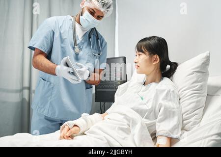 Un médecin méconnaissable portant un masque démontrant quelque chose à un jeune patient asiatique sur un écran de tablette numérique Banque D'Images