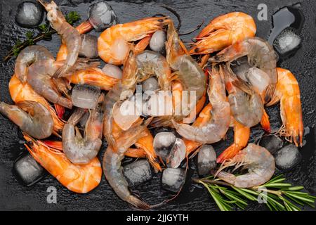 Crevettes crues et glace sur une surface en pierre humide. Branche de romarin. Pose à plat. Arrière-plan noir Banque D'Images