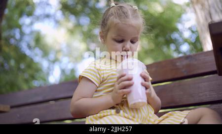 Une petite fille boit un milkshake à travers une paille. Portrait en gros plan d'une petite fille mignonne assise sur le banc du parc et buvant du milk-shake Banque D'Images