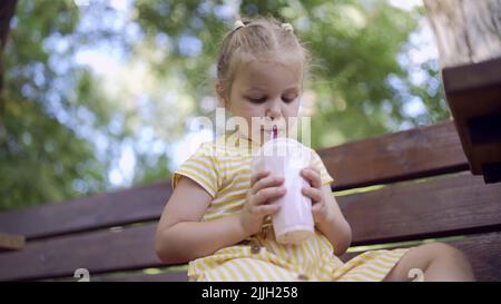 Une petite fille boit un milkshake à travers une paille. Portrait en gros plan d'une petite fille mignonne assise sur le banc du parc et buvant du milk-shake Banque D'Images