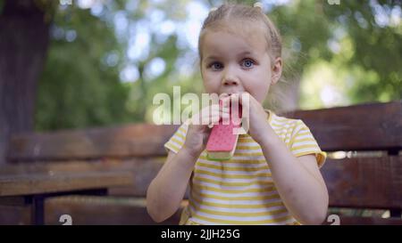 Gros plan de la jolie petite fille mange de la glace. Gros plan d'une fillette assise sur le banc du parc et mangeant de la glace. Banque D'Images