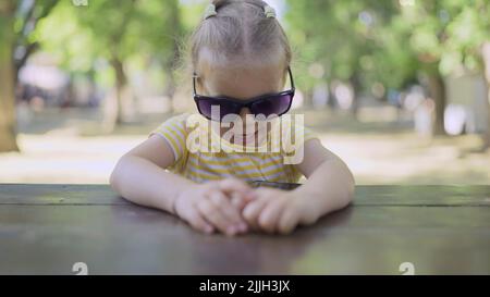 La petite fille dans les lunettes de soleil de maman apprend la langue étrangère en répétant des mots à partir de téléphone mobile. Portrait en gros plan de l'enfant fille assis dans la ville par Banque D'Images