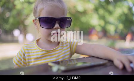 La petite fille dans les lunettes de soleil de maman apprend la langue étrangère en répétant des mots à partir de téléphone mobile. Portrait en gros plan de l'enfant fille assis dans la ville par Banque D'Images