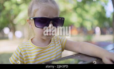 La petite fille dans les lunettes de soleil de maman apprend la langue étrangère en répétant des mots à partir de téléphone mobile. Portrait en gros plan de l'enfant fille assis dans la ville par Banque D'Images