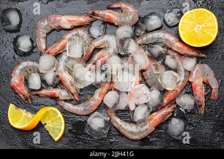 Crevettes et tranches de citron parmi la glace. Planche à pierres humide. Pose à plat. Arrière-plan noir Banque D'Images