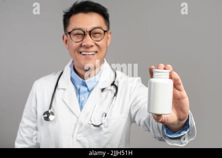Sourire homme asiatique mature médecin en manteau, verres, montre bocal de médicaments isolés sur fond gris, gros plan Banque D'Images