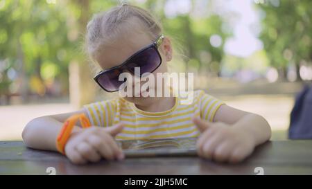 Odessa, Ukraine, Europe de l'est. 26th juillet 2022. La petite fille dans les lunettes de soleil de maman apprend la langue étrangère en répétant des mots à partir de téléphone mobile. Portrait en gros plan d'une jeune fille assise dans un parc municipal et apprenant la langue étrangère à l'aide d'un téléphone portable (Credit image: © Andrey Nekrasov/ZUMA Press Wire) Banque D'Images