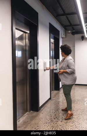 Vue latérale d'une femme d'affaires biraciale de taille moyenne, appuyant sur le bouton-poussoir de l'ascenseur dans le bâtiment de bureau Banque D'Images