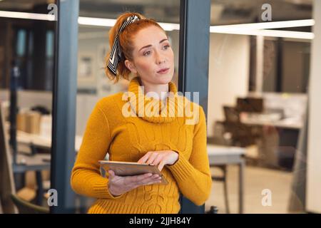 Femme d'affaires caucasienne attentionnée regardant loin tout en se tenant debout avec une tablette numérique au bureau Banque D'Images