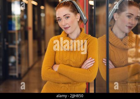 Portrait d'une femme d'affaires caucasienne souriante avec les bras croisés s'inclinant sur le mur de verre au bureau Banque D'Images