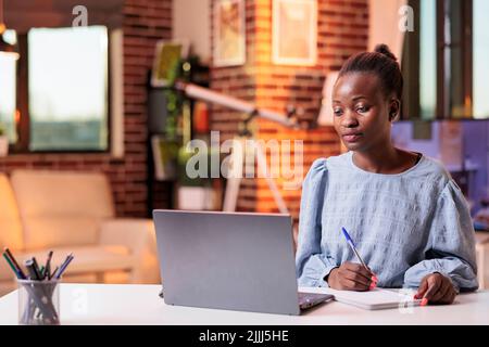 Jeune femme étudiant, utilisant un ordinateur portable et prenant des notes dans le bloc-notes. Étudiante africaine américaine à distance fréquentant des cours à distance et apprenant avec des cours en ligne sur Internet Banque D'Images