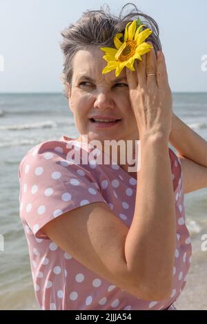 Femme mariée aux cheveux gris dans une robe rose avec des pois blancs tenant la fleur jaune dans sa main levée regardant loin. Concept de beaty naturel. Banque D'Images