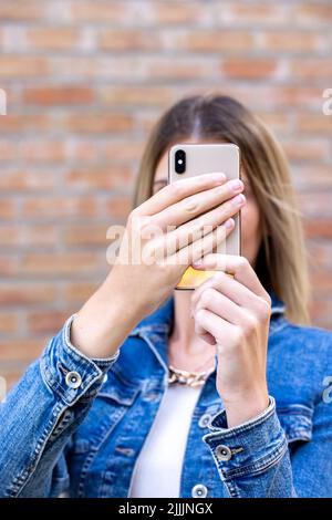 Photo de gai mignon charmant joli joli beau joli joli joli joli petit-fille blond, pâle à la peau prenant selfie avec son téléphone tenu dans ses mains portant Jean veste isolée sur fond de brique de couleur rouge. Photo de haute qualité Banque D'Images