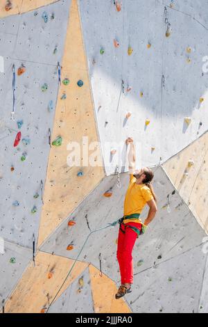 Le grimpeur sportif est suspendu d'une main sur un mur d'escalade artificiel à l'extérieur. L'homme a poussé ses mains dans un sac de poudre de magnésie derrière son dos. Banque D'Images