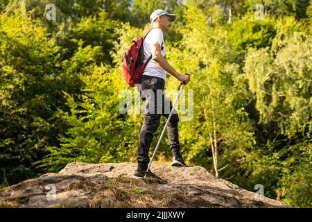 Jeune randonneur avec sac à dos et bâtons de randonnée debout sur le bord de la falaise et regardant les montagnes en été en plein air. Banque D'Images