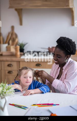 Une femme souriante à la peau foncée qui se porte aux cheveux de la jolie fille avec le syndrome de Down Banque D'Images