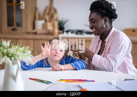 Une femme souriante à la peau foncée qui se porte aux cheveux de la jolie fille avec le syndrome de Down Banque D'Images