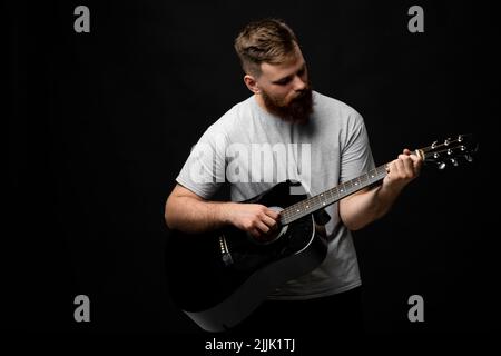 Homme barbu concentré et brutal dans un t-shirt gris jouant de la guitare acoustique. Portrait d'un chanteur et guitariste masculin. Banque D'Images