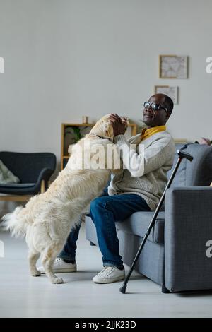 Homme africain aveugle en lunettes jouant avec le chien guide assis sur le canapé à la maison Banque D'Images