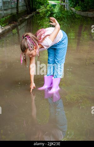 Une fille joyeuse tressée dans des bottes en caoutchouc joue dans des flaques boueuses. Banque D'Images