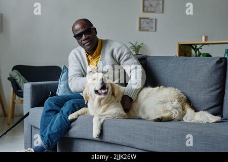 Portrait d'un homme aveugle africain dans des lunettes sombres reposant sur un canapé dans le salon avec son chien-guide Banque D'Images