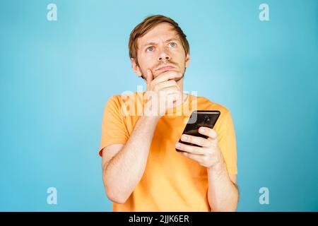 Portrait d'un homme pensif attentionné tenant un smartphone d'une main, touchant le menton sur un fond bleu. Banque D'Images